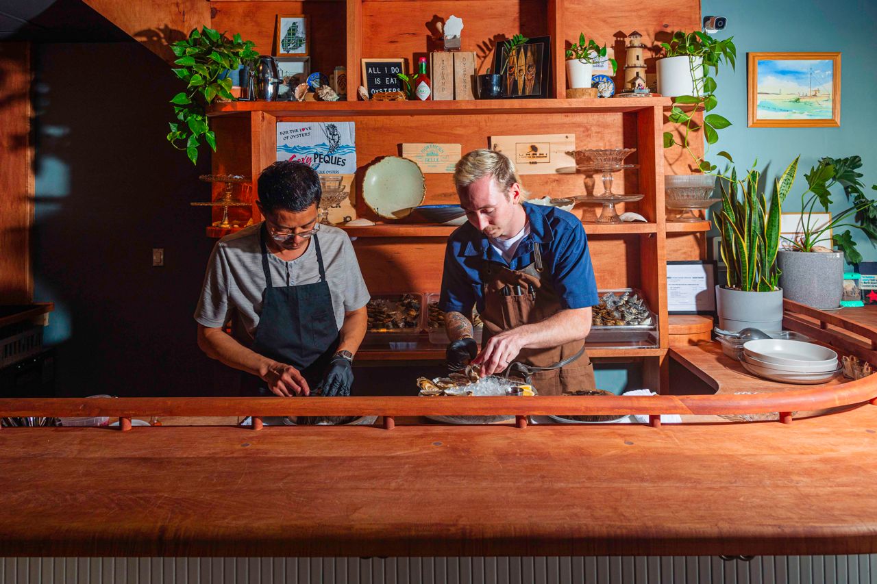 Two men shuck oysters behind a bar